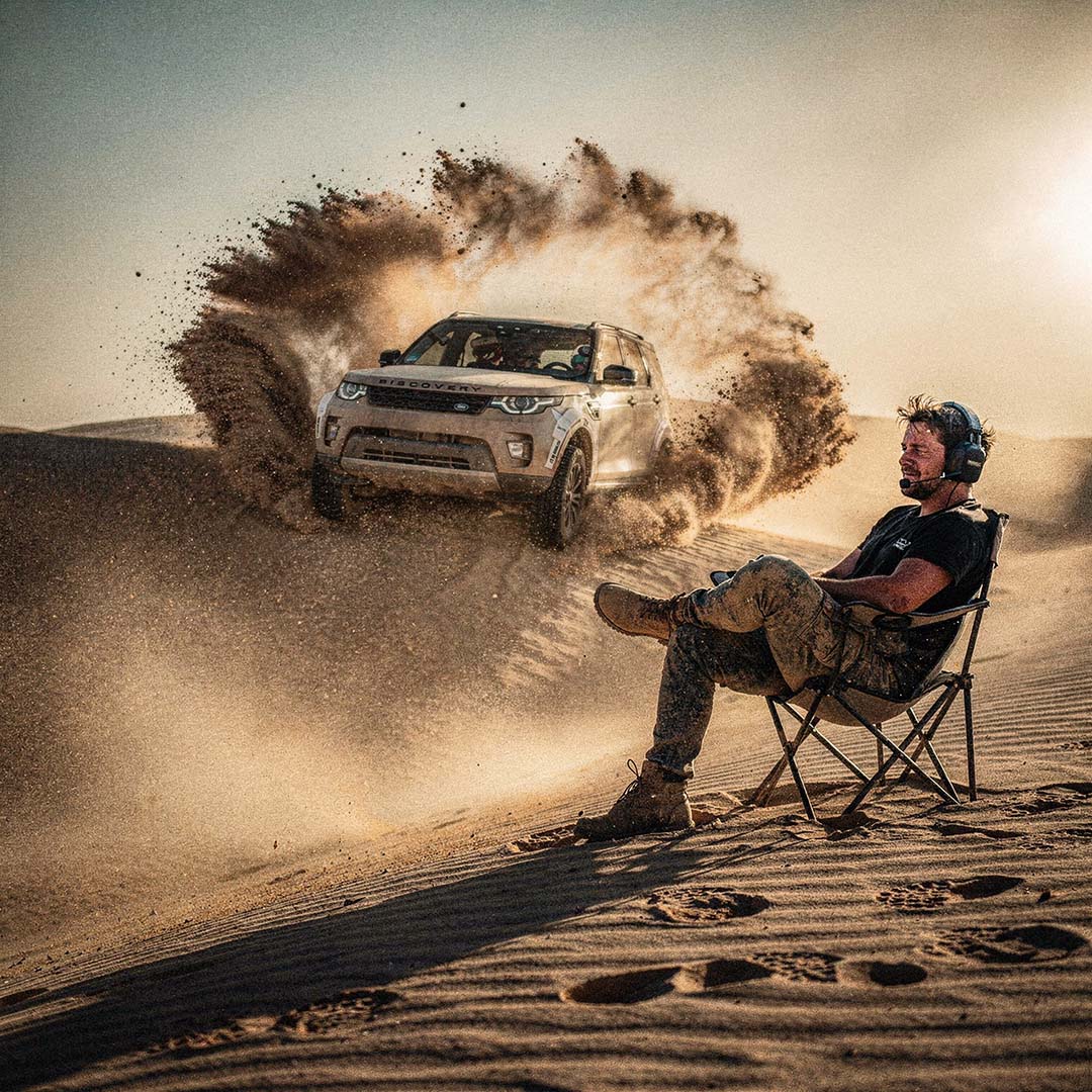 Hartscharfer 1:1 Action-Shot: Ein schwarzer Land Rover Discovery springt über eine Düne mit massiver Sandwolke, im Vordergrund ein entspannter Reporter mit Headset, Art Direction von The Impression.