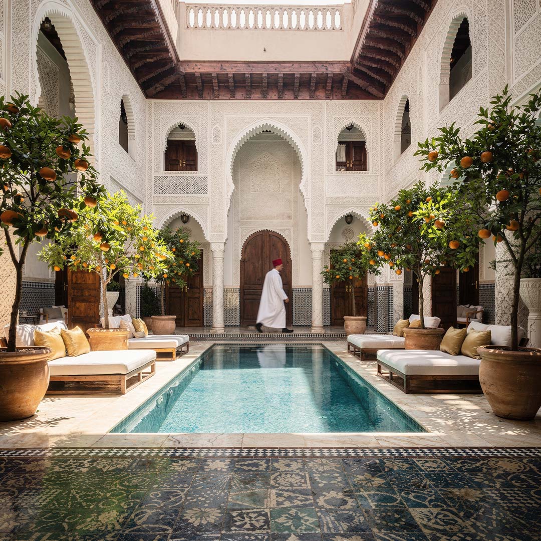Hartscharfer 1:1 Architektur-Shot: Ein heller marokkanischer Riad mit einem Kellner in weißer Djellaba im Motion Blur im Hintergrund, lebendiger Luxus und präzise Art Direction von The Impression.