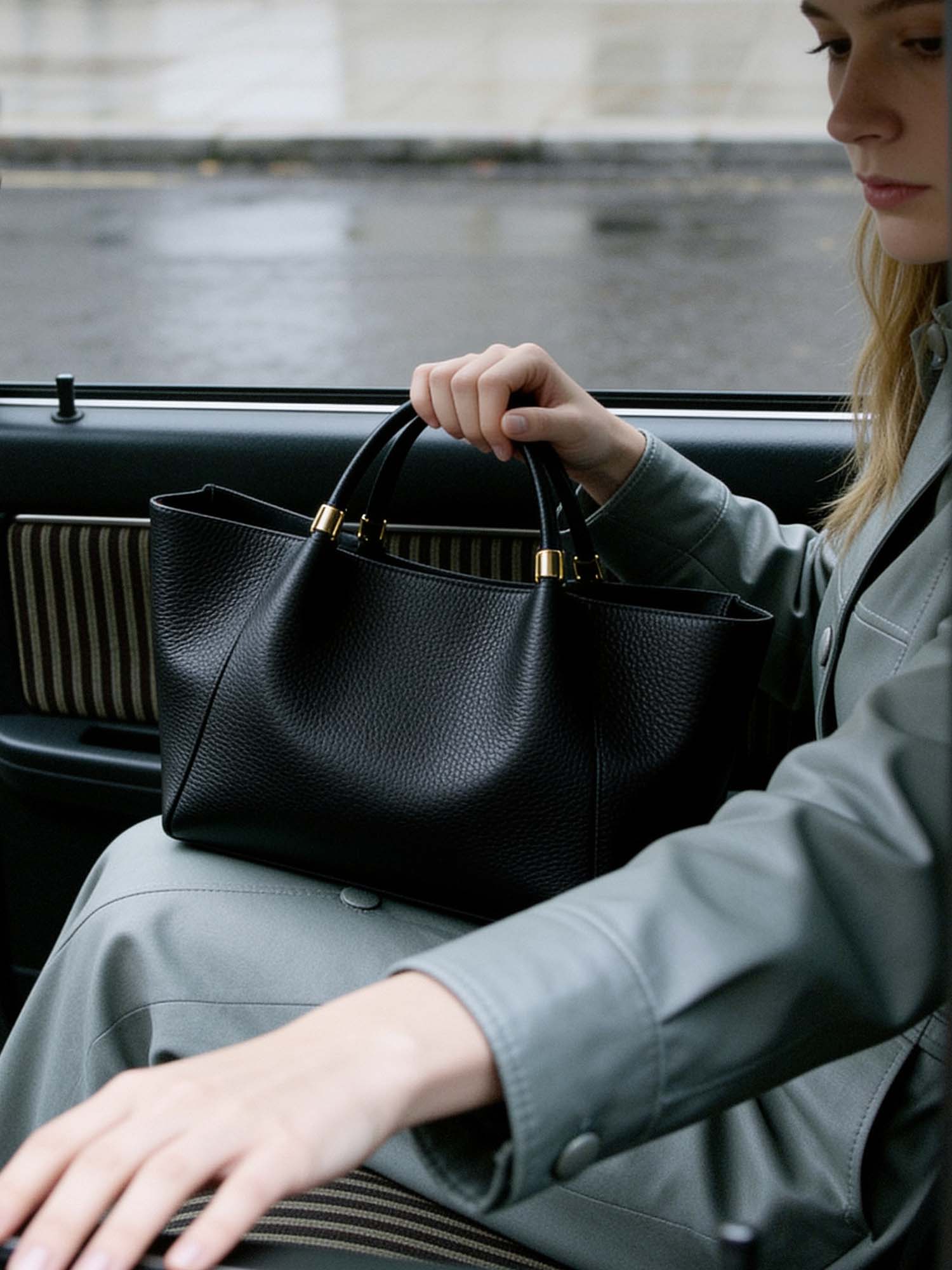 A taxi idles at a city curb in flat neutral daylight. A woman in the back seat, seen from outside through the open door: a black pebbled-leather tote sits on her lap