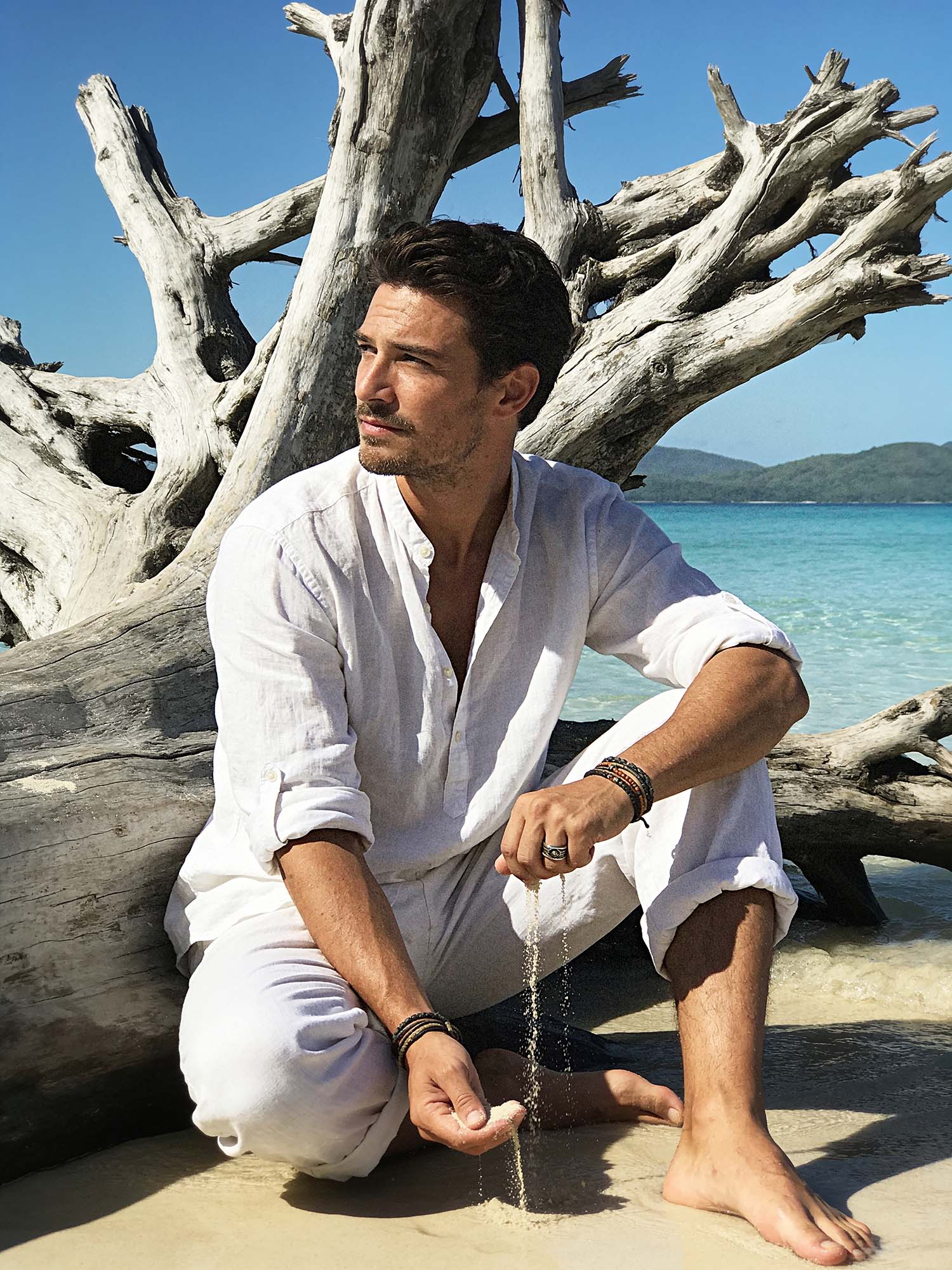 A man sits on a sunny sand beach at the waterline, barefoot, in profile to the viewer; torso leaned slightly forward; gaze thoughtfully to the left, toward the horizon, not at the camera.