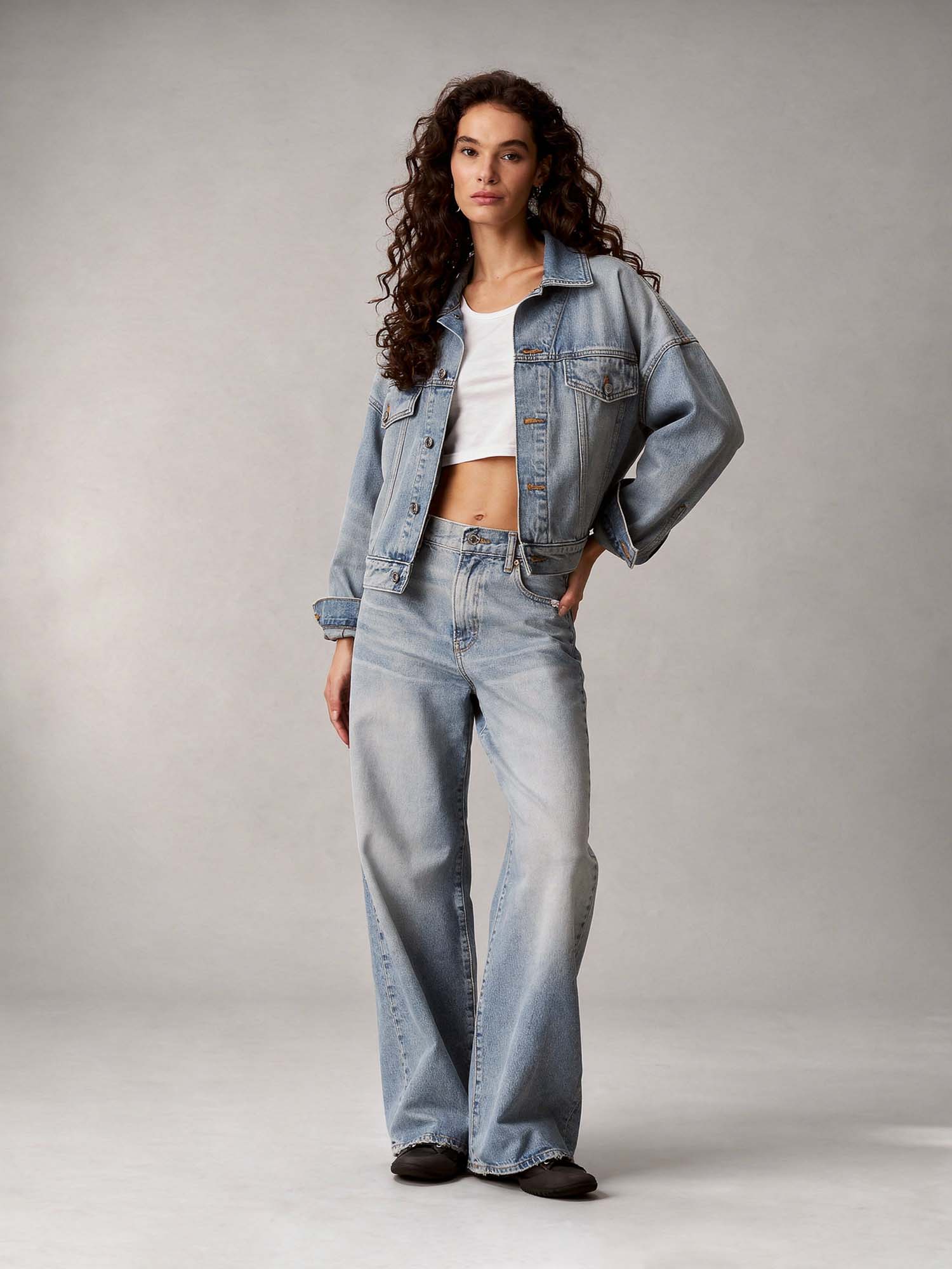 A full-length portrait in a professional studio. A young woman with long, dark, curly hair, wearing a short white t-shirt that shows her midriff, an open light-blue denim jacket with rolled cuffs, and baggy light-blue denim jeans