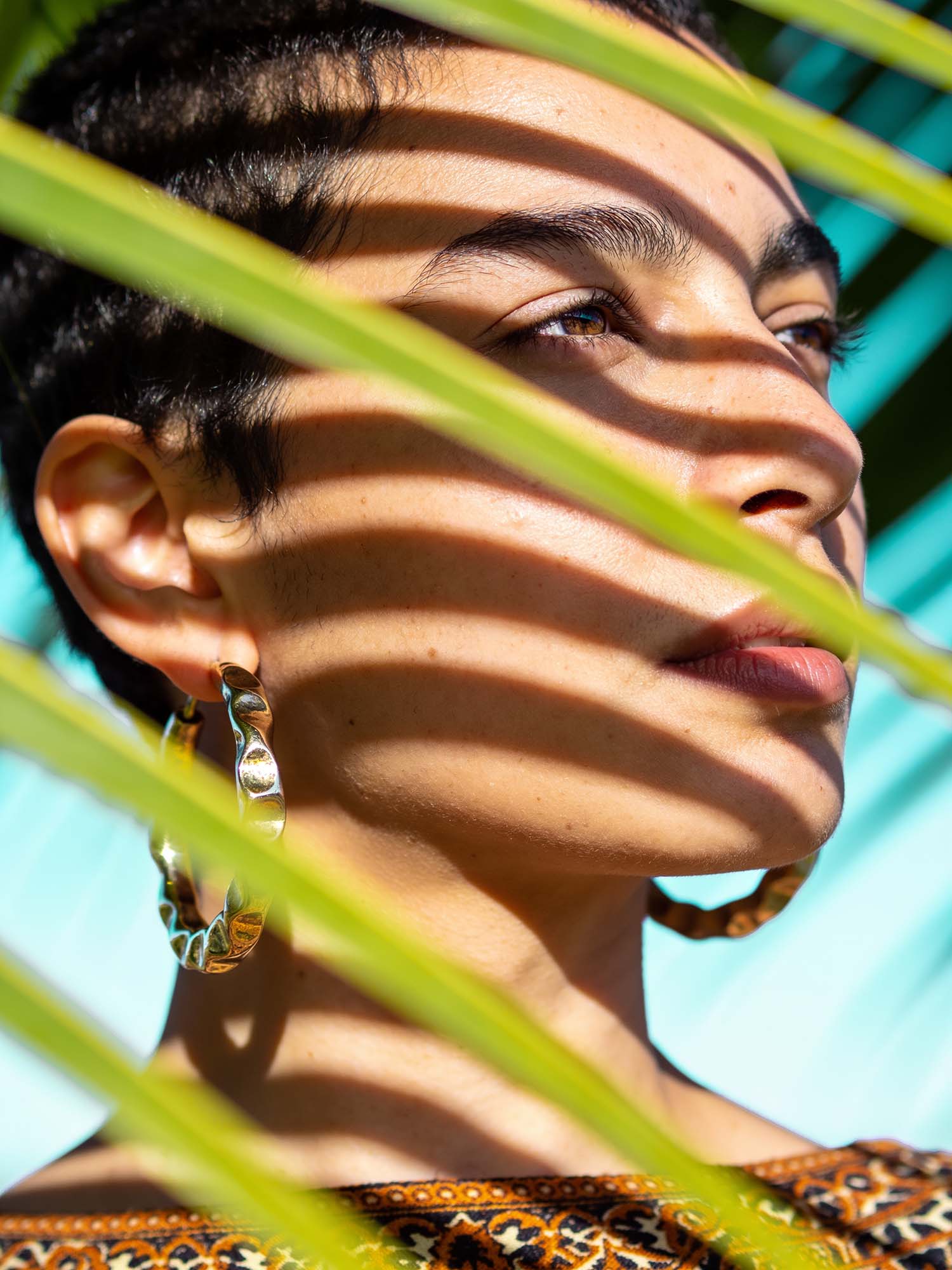 A model's head is turned slightly to the right of the viewer, gazing through palm leaves on a sunny day