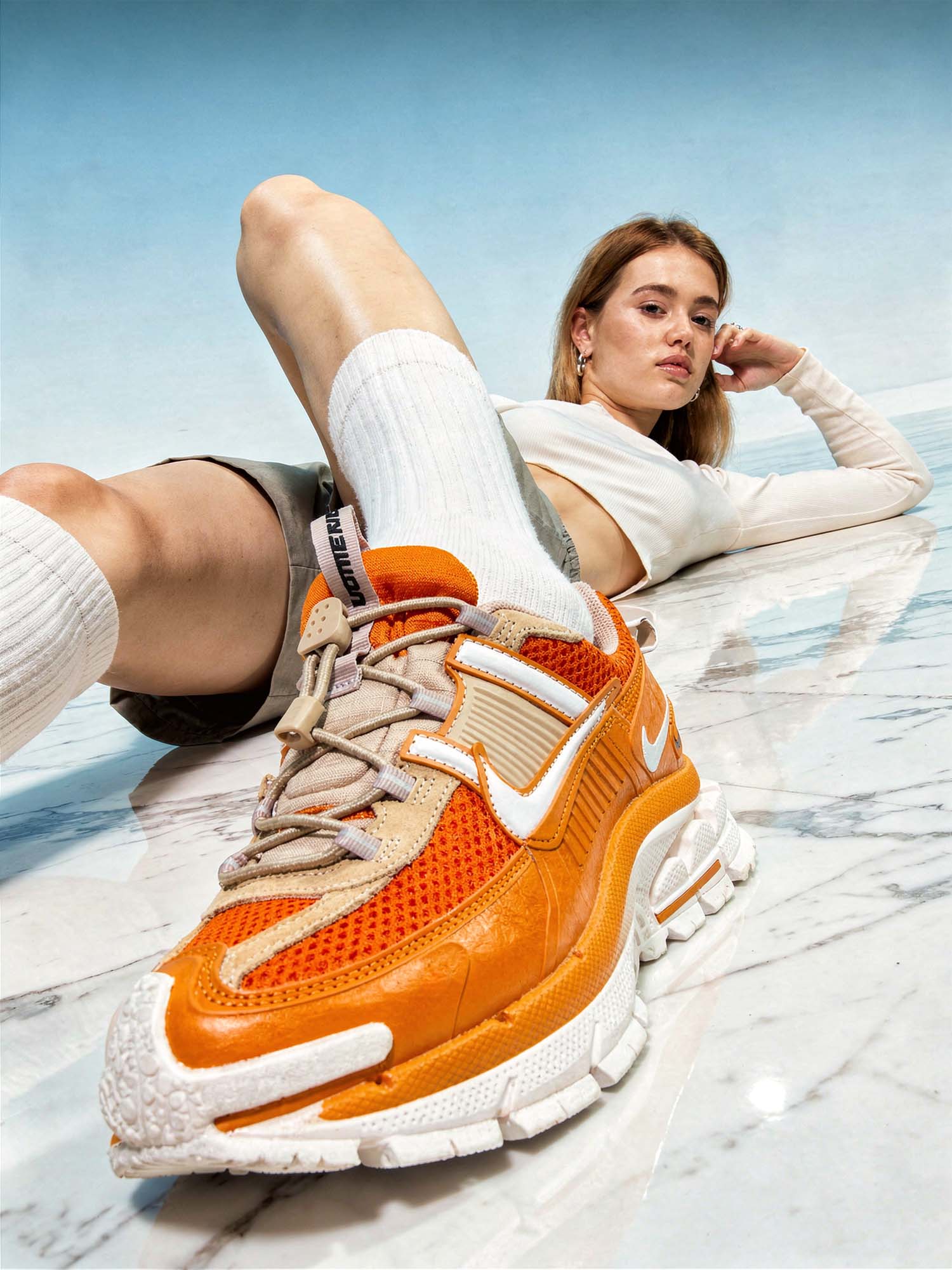 Fashion studo product photography. A young European woman with fair skin, light brown hair, lies on a highly polished dark stone floor. Her right leg is raised with her right foot in the extreme foreground