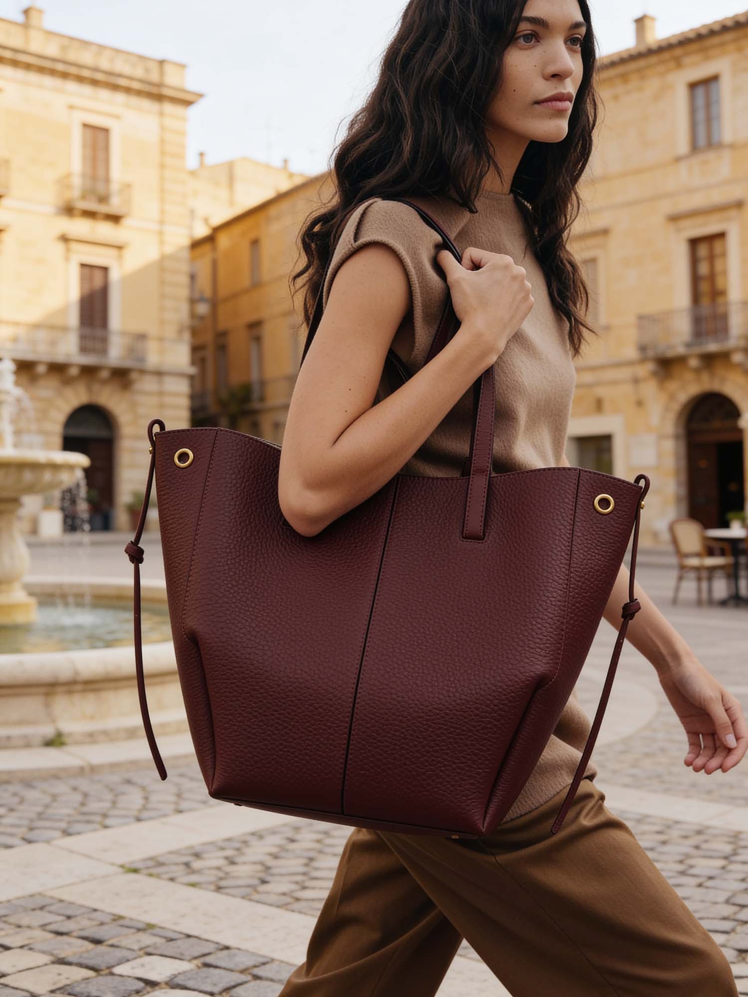 A woman walks along a quiet country road in the Sicilian countryside, seen from behind, mid-stride. She carries the crescent hobo bag in her right hand, swinging easily at her side