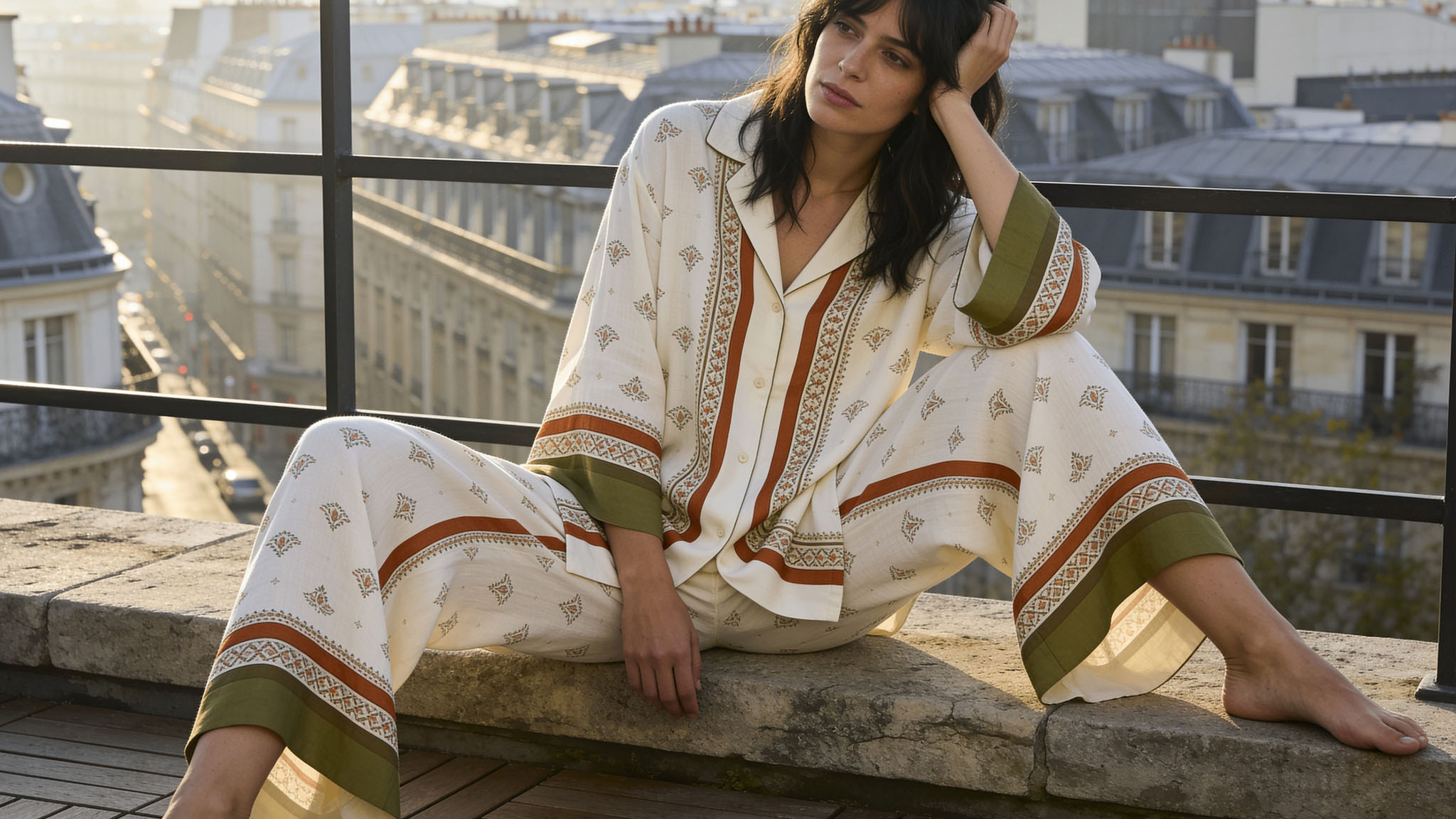 An adult fashion model with natural posture sits on a low stone wall of a Parisian penthouse rooftop patio overlooking the city, her head resting on her hand, her expression distant and reflective.