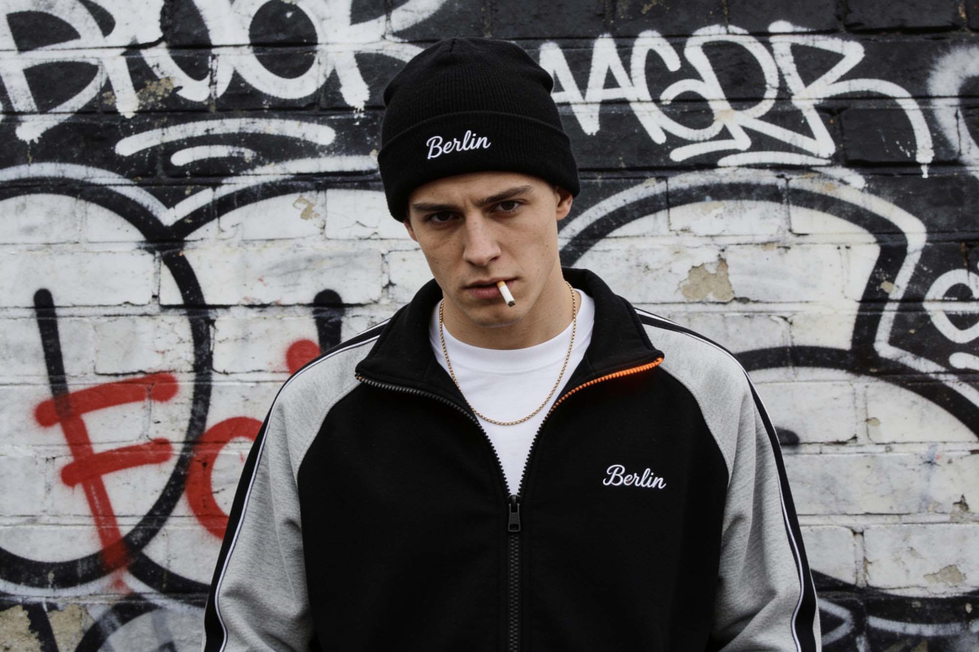A young man in his early 20s, standing in front of a brick wall covered in layered graffiti tags, Berlin. He wears a black beanie, a black and grey zip-up jacket over a white t-shirt, and a thin gold chain necklace.